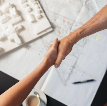 MitarbeiterInnen von Swietelsky reichen sich die Hand, Grundrissplan und Kaffeetasse im Hintergrund